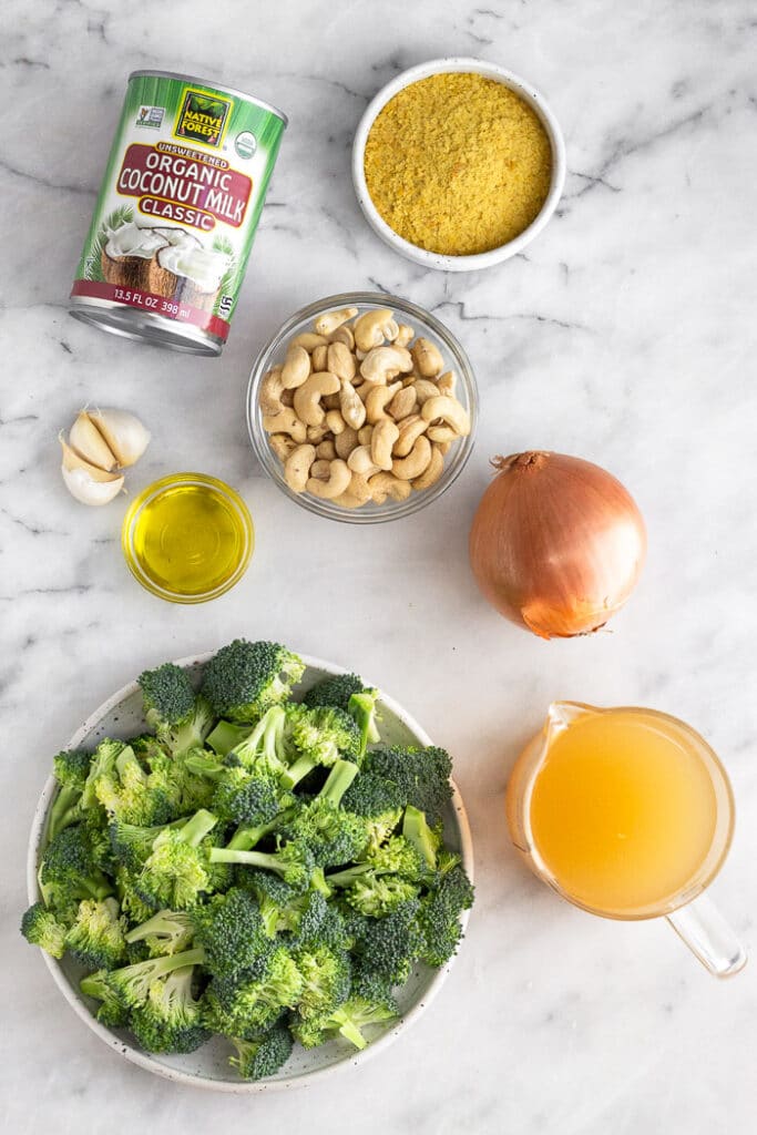Marble countertop with a can of coconut milk, bowl of nutritional yeast, bowl of cashews, yellow onion, measuring cup of vegetable broth, plate of broccoli florets, small bowl of oil, and 3 cloves of garlic.