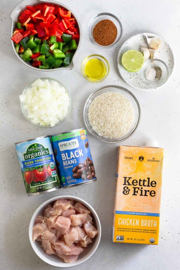 White counter with a bowl of taco seasoning, a plate of half a lime, 3 cloves of garlic, and a small bowl of salt and pepper, a bowl of olive oil, a bowl of uncooked white rice, a carton of chicken broth, a bowl of cubed raw chicken, a can of canned tomatoes, a can of black beans, a bowl of diced white onion, and a bowl of diced bell peppers.