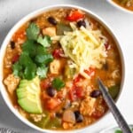 A bowl of chicken fajita soup topped with sliced avocado, cilantro, and shredded cheese. Around it is another bowl of soup, a bowl of tortilla chips, a striped kitchen towel, and a plate of sliced avocado and cilantro.