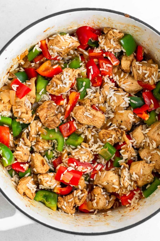 Large white dutch oven filled with seasoned cubed chicken breasts, diced bell peppers, and uncooked white rice all mixed together.