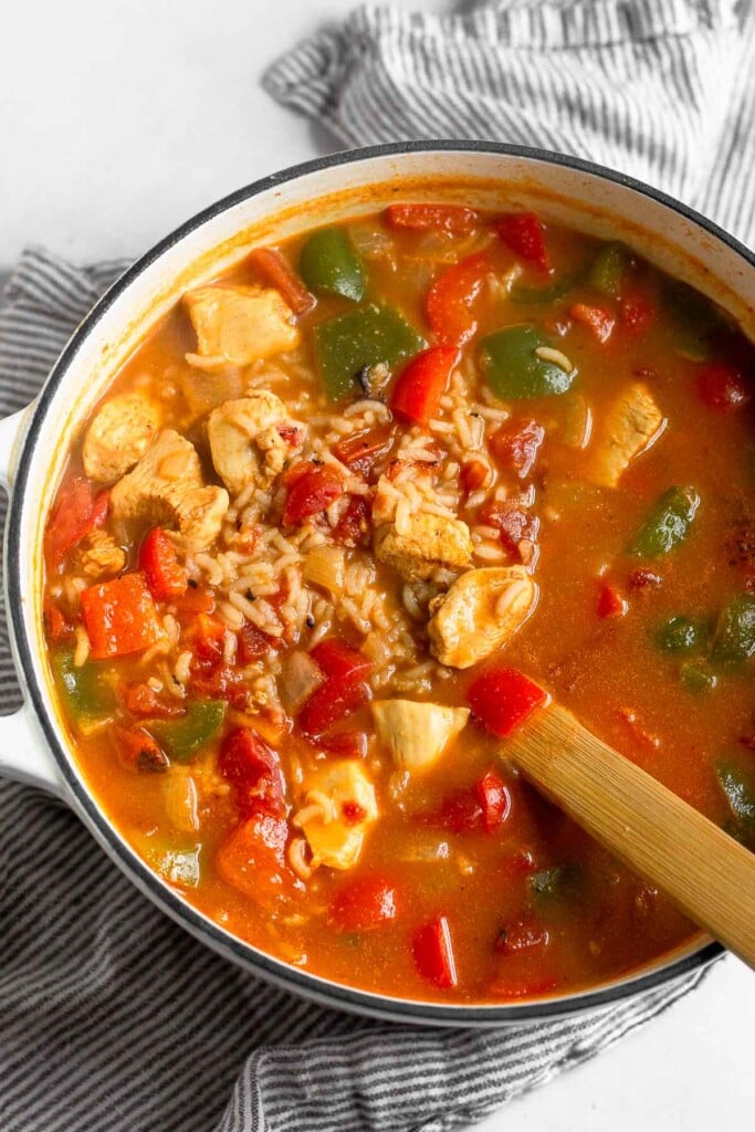 Large white dutch oven filled with chicken and rice soup with veggies. A wooden spoon is sticking out of it.