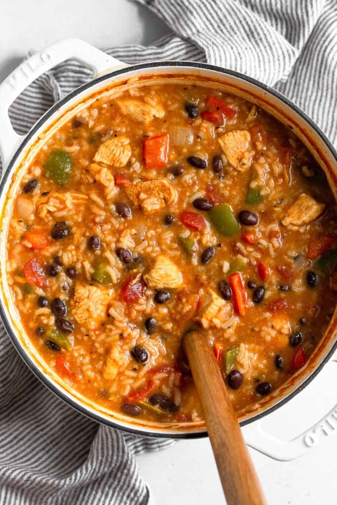 Large pot of chicken soup with rice, black beans, and peppers with a wooden spoon in it. The pot is sitting on a striped kitchen towel.