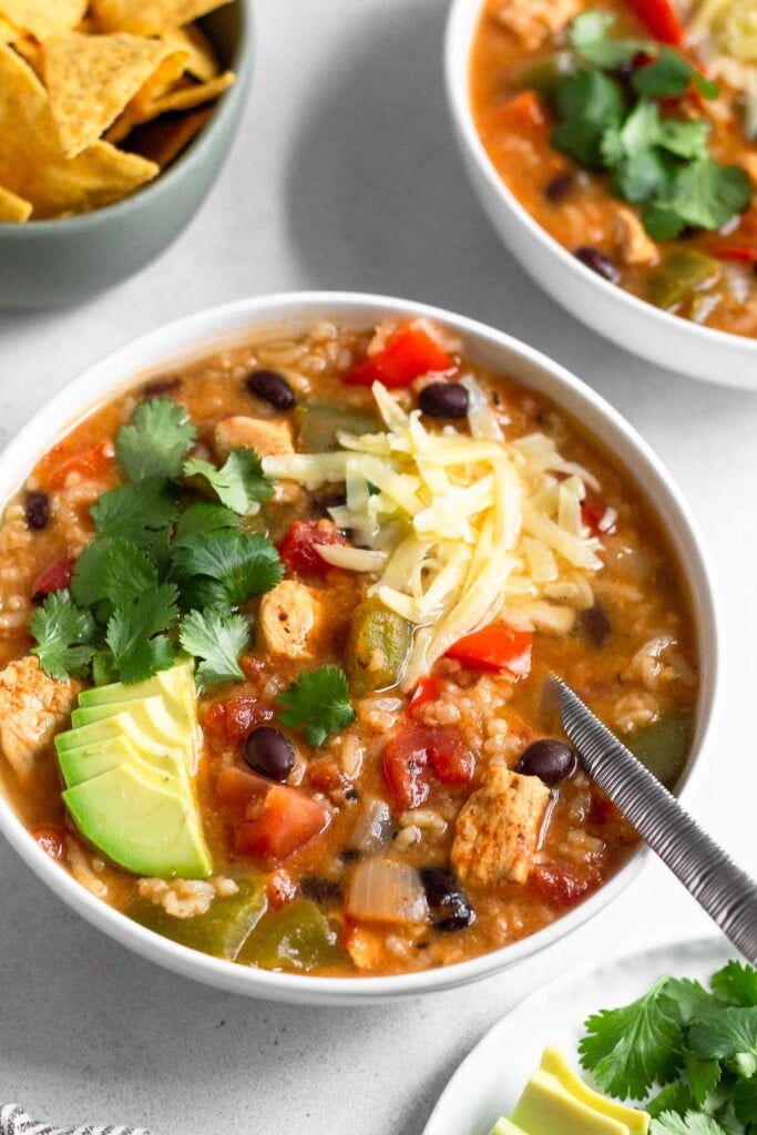 White bowl with fajita chicken soup topped with sliced avocado, cilantro, and shredded cheese. A spoon is in the soup. Around the bowl of soup is a bowl of tortilla chips, another bowl of soup, and a plate of avocado and cilantro.