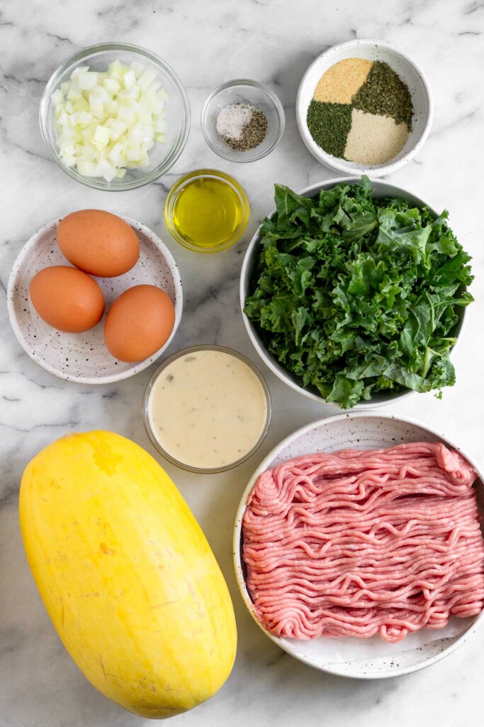 White marble counter with a bowl of spices, a bowl of chopped kale, a bowl of raw ground turkey, a whole spaghetti squash, a bowl of ranch, a plate of 3 brown eggs, a bowl of olive oil, a bowl salt and pepper, and a bowl of diced onion.