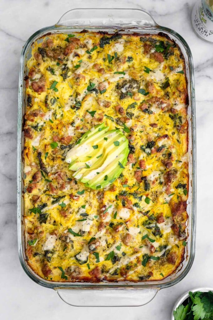 Overhead shot of spaghetti squash and ground turkey casserole in a large casserole dish. It is topped with sliced avocado, ranch dressing, and parsley. Next to it is a bowl of parsley and a bottle of ranch dressing.