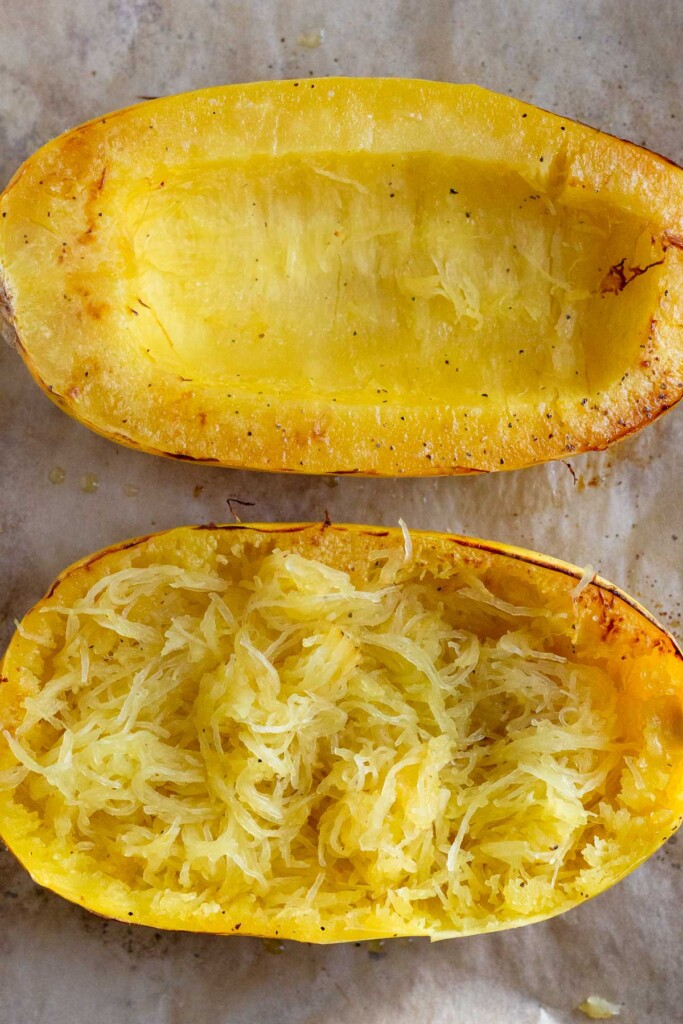 Baking sheet lined with parchment paper with two halves of spaghetti squash that have been baked. One of the halves has been threaded.