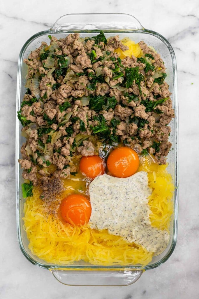 Casserole dish with cooked spaghetti squash, ranch dressing, three raw eggs, and a cooked meat and veggie mixture before it is mixed together.