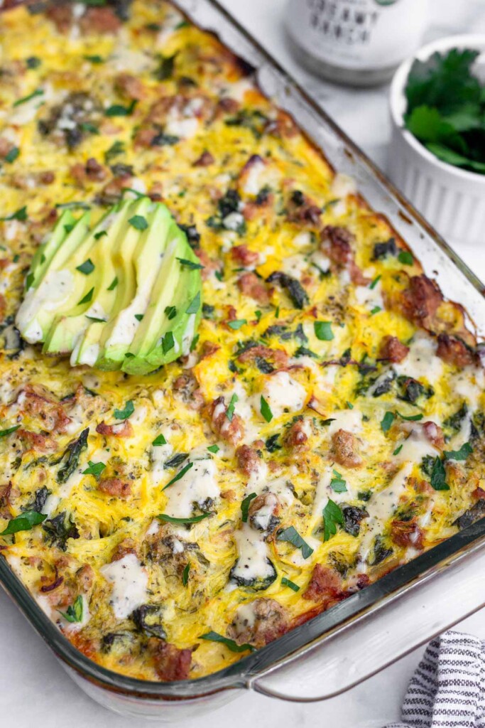 Ground turkey casserole with spaghetti squash. It is topped with ranch dressing, parsley, and sliced avocado. Behind the pan is a bowl of parsley and a bottle of ranch.
