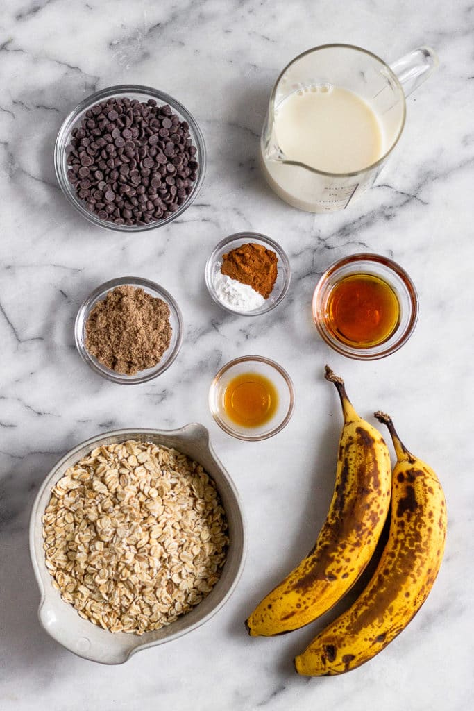 White counter top with a bowl of chocolate chips, glass of milk, bowl of maple syrup, 2 brown bananas, bowl of rolled oats, bowl of vanilla extract, bowl of flaxseed, and bowl of spices.