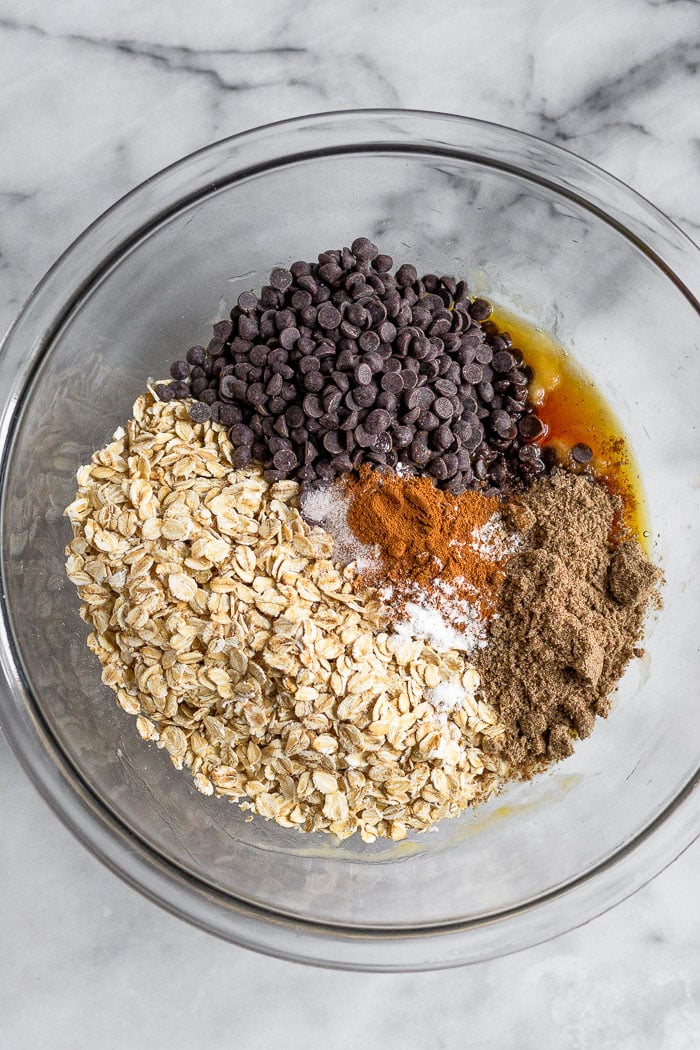 Glass bowl filled with rolled oats, mini chocolate chips, ground flaxseed, maple syrup, vanilla extract, cinnamon, baking soda, and salt.