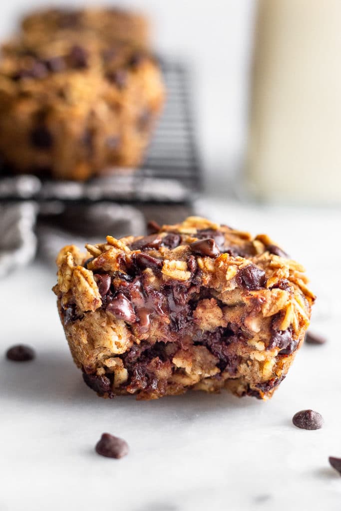 Healthy oatmeal chocolate chip muffin with a bite taken out of it. Behind it is a rack of more muffins and a jar of milk.