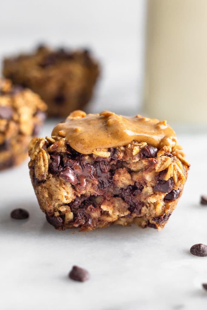 Healthy oatmeal muffin topped with peanut butter with a bite taken out of it. Behind it is some more cups and a glass of milk.