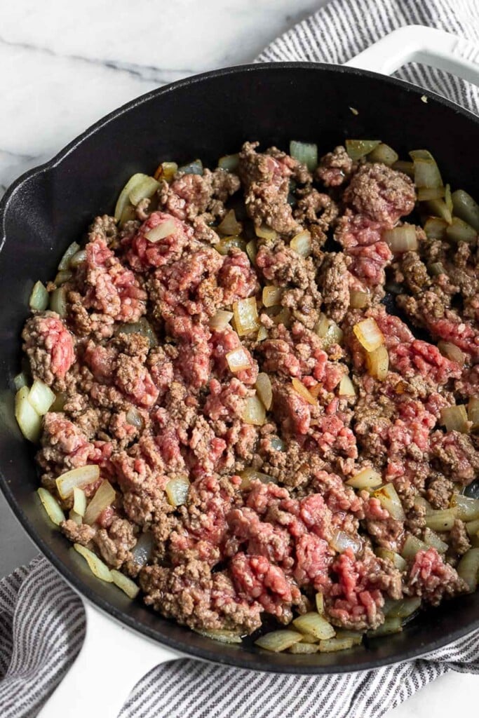 Cast iron filled with sautéed onion and ground beef that is halfway cooked.