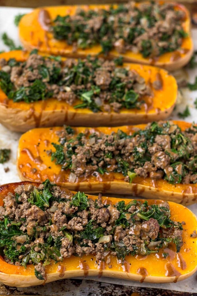 Large baking sheet filled with four kale and beef stuffed butternut squash halves drizzled with tahini sauce.