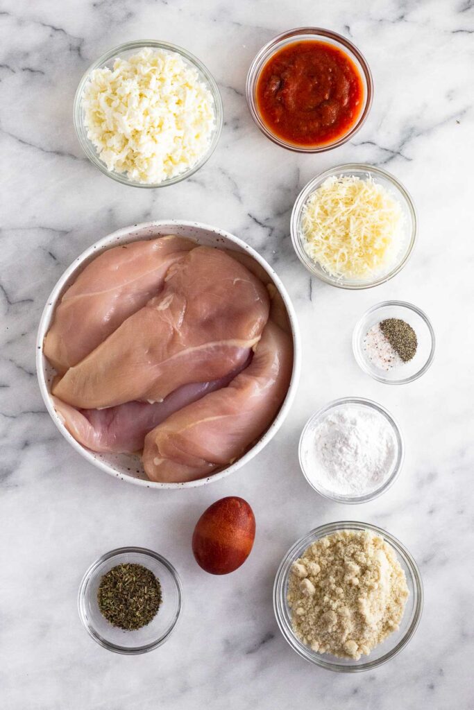 White counter with a bowl of shredded cheese, bowl of marinara sauce, another bowl of shredded cheese, bowl of salt and pepper, two bowls of flour, an egg, bowl of spices, and a bowl of raw chicken breasts.