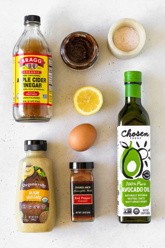 White counter with a bowl of salt, a bottle of avocado oil, a jar of cayenne pepper, a bottle of dijon mustard, a brown egg, half a lemon, a jar of apple cider vinegar, and a jar of chipotle paste.