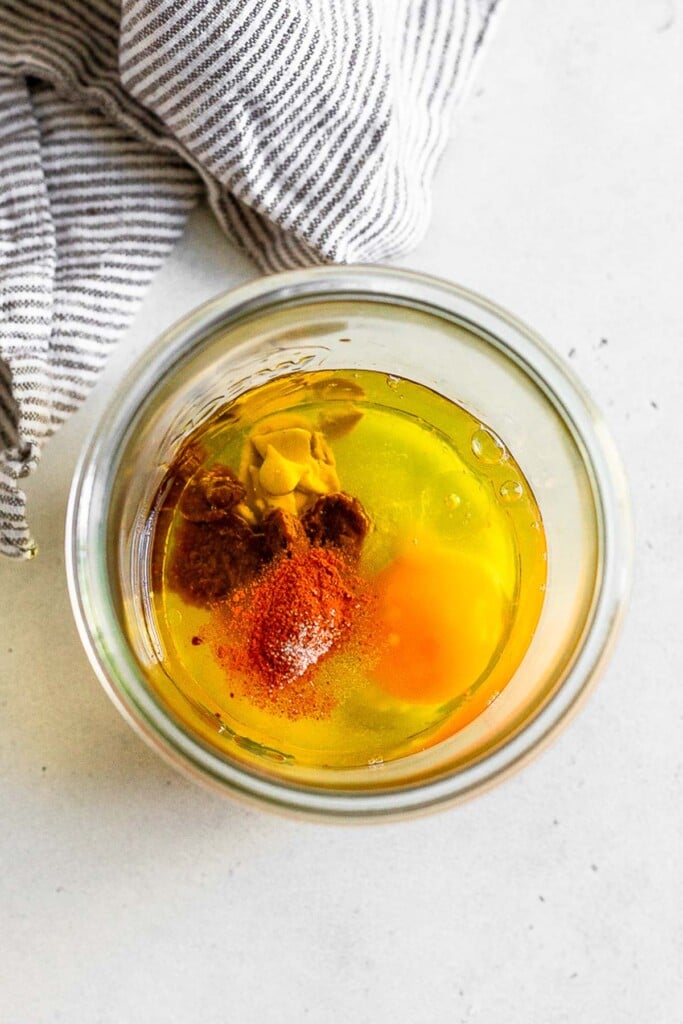 Overhead shot of a jar filled with a raw egg, oil, mustard, and some spices. Next to it is a striped kitchen towel.