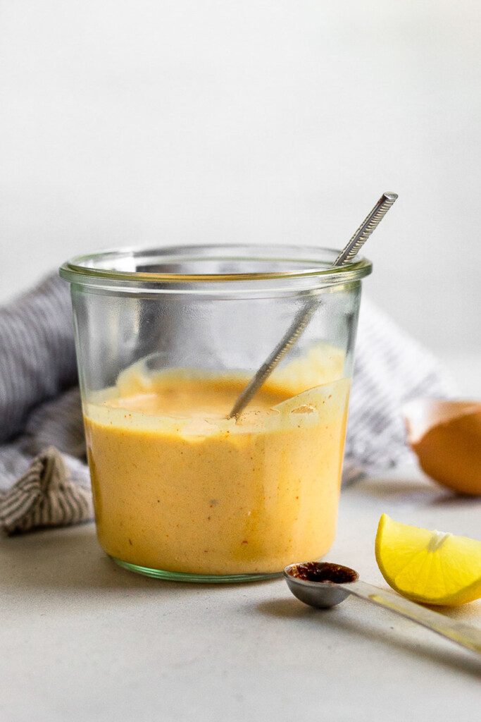 Jar with homemade spicy mayo with a spoon sticking out of it. Next to it is a small teaspoon, lemon wedge, egg shell, and a striped towel.