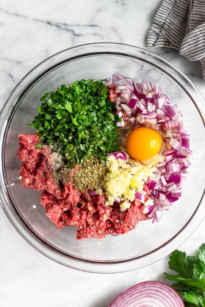 Large glass bowl of ground lamb, diced red onion, chopped fresh herbs, egg, minced garlic, spices, and salt and pepper.
