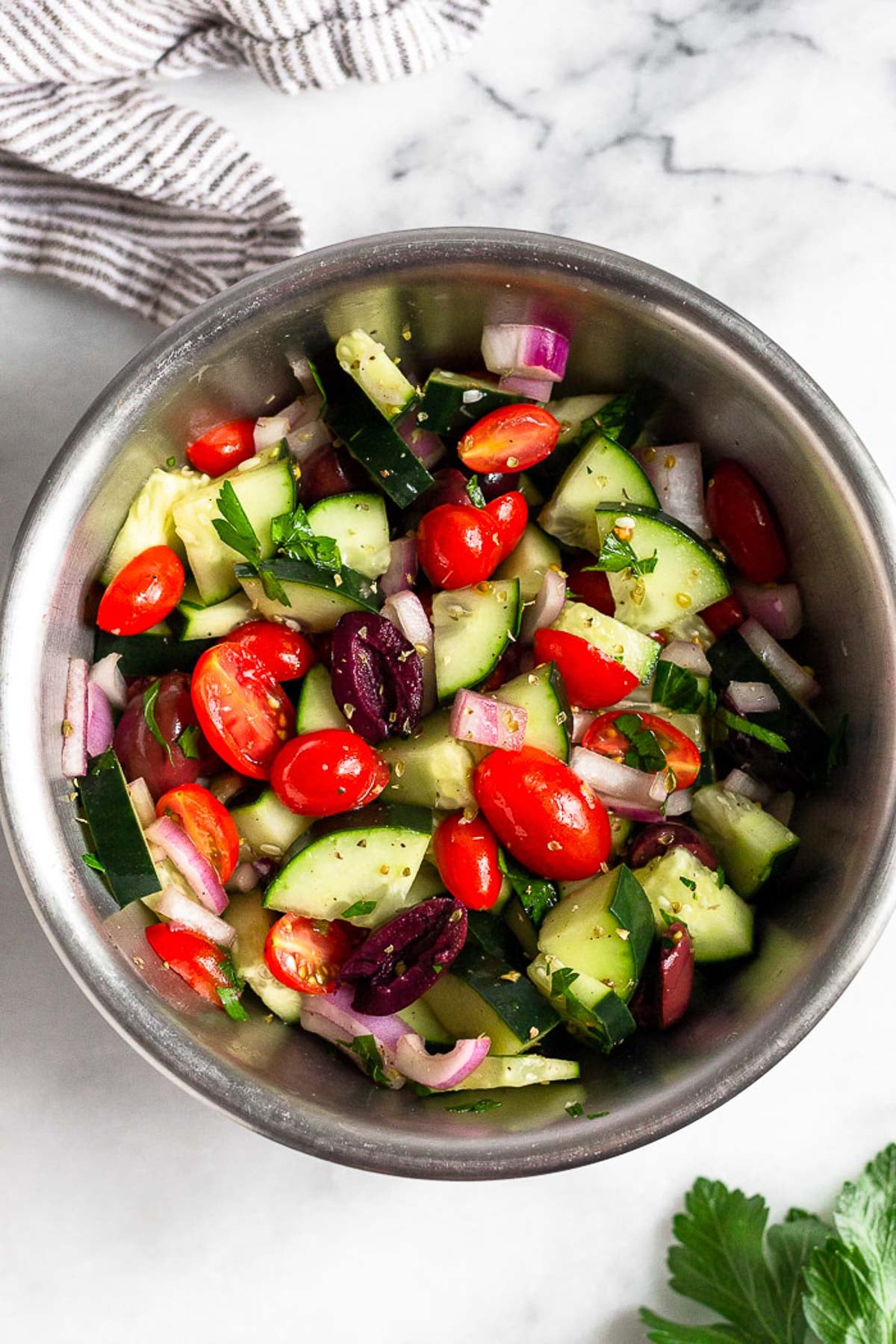 Bowl of chopped cucumber, tomatoes, olives, red onion, and parsley coated in dressing.