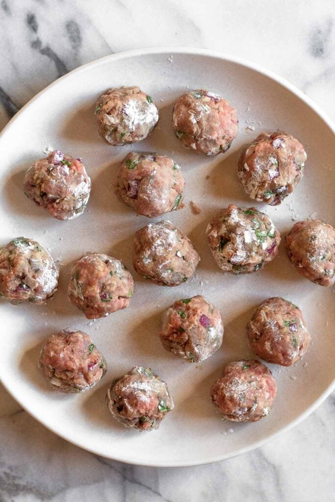 Raw greek meatballs coated in flour on a large white plate.