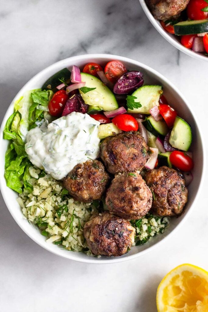 Greek bowl with lamb meatballs, cauliflower rice, romaine, greek salad, and tzatziki sauce. Around it is another bowl and half a lemon wedge.