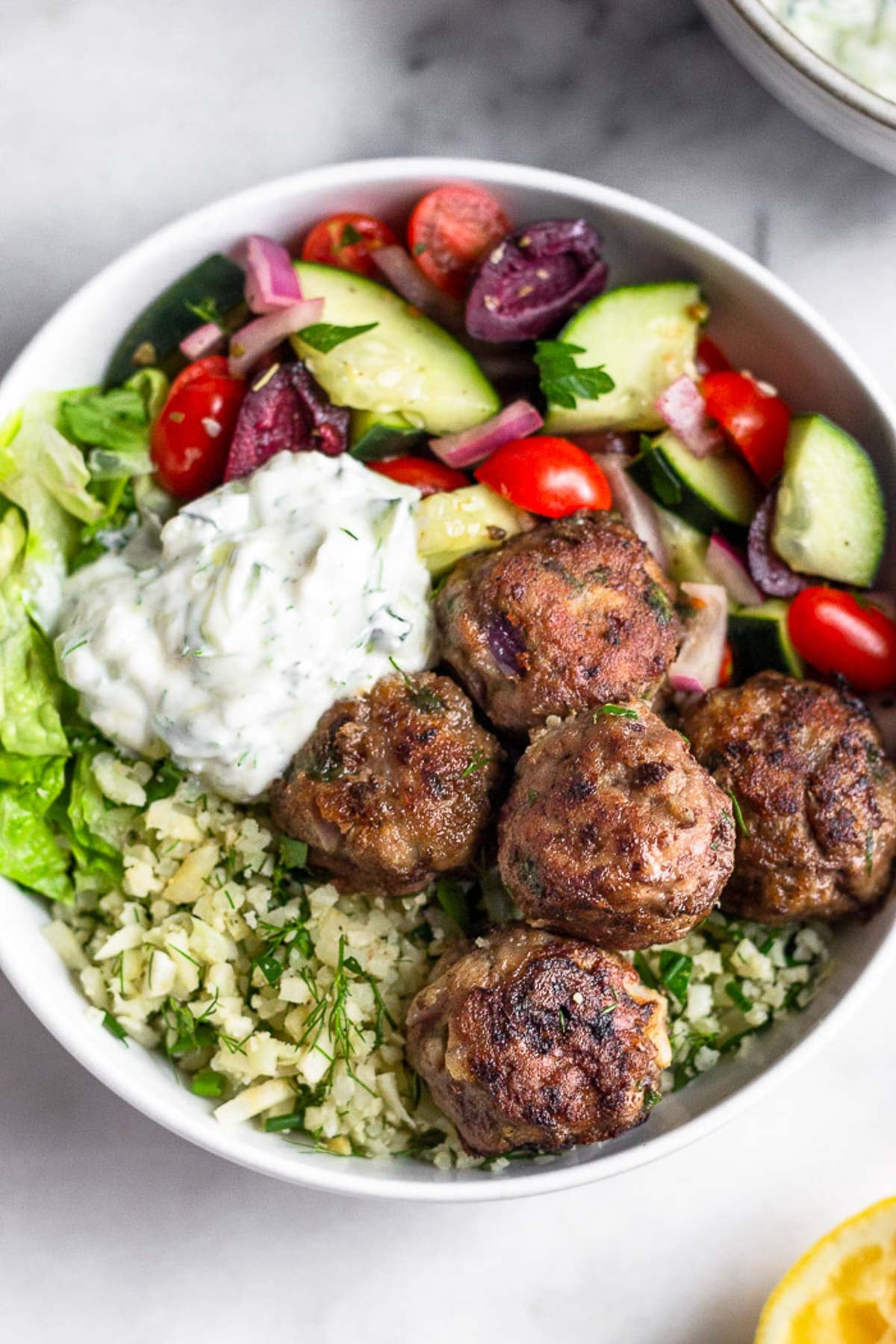 Overhead shot of a greek bowl with greek lamb meatballs, herby cauliflower rice, chopped romaine, greek salad, and tzatziki sauce.