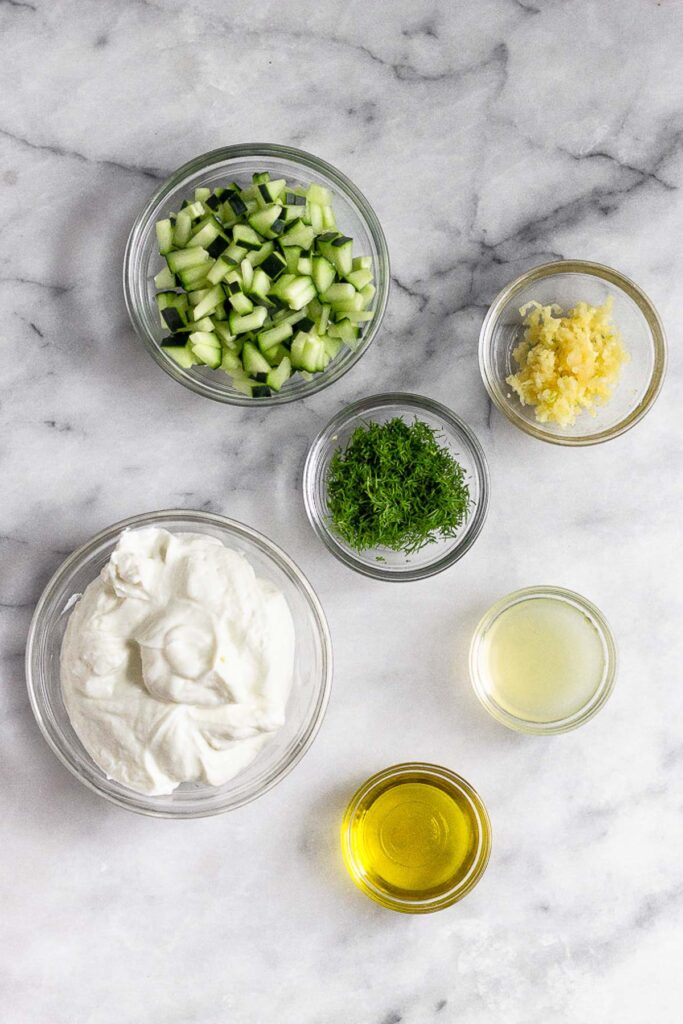 Marble counter with a bowl of diced cucumber, a bowl of minced garlic, a bowl of fresh dill, a bowl of lemon juice, a bowl of olive oil, and a bowl of greek yogurt.