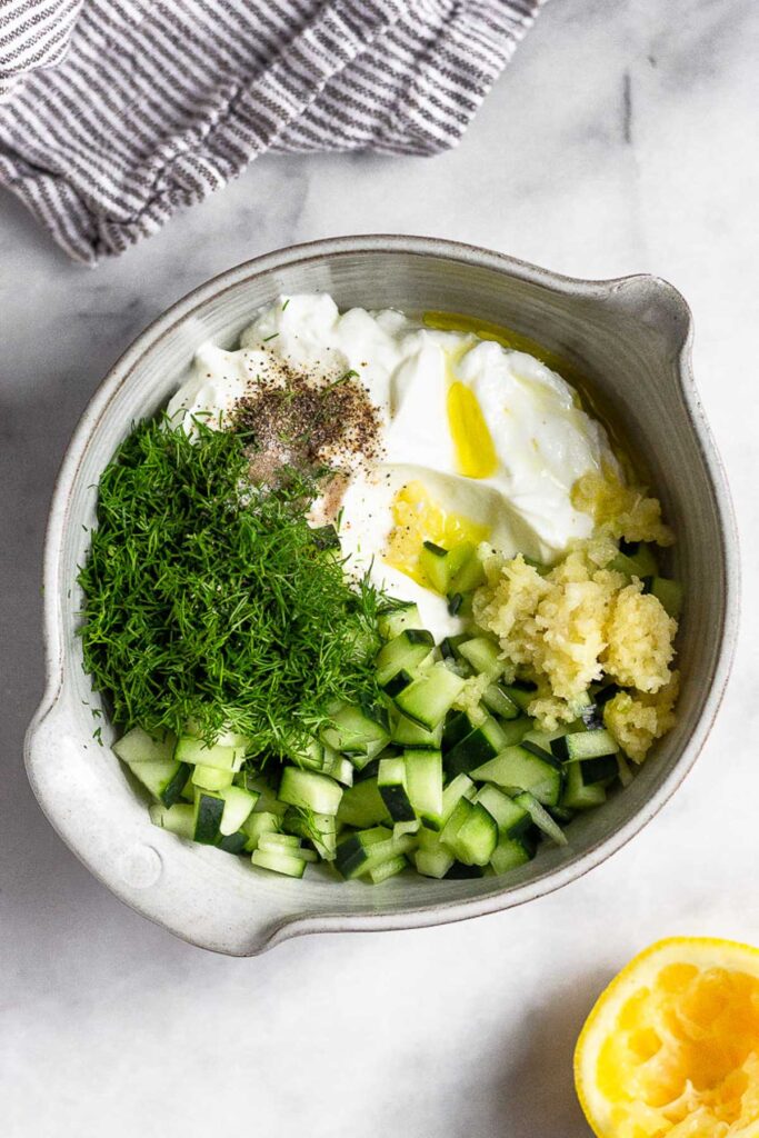 Bowl filled with greek yogurt, chopped cucumber, fresh dill, minced garlic, lemon juice, olive oil, and salt and pepper. Next to it is a kitchen towel and half a juiced lemon.