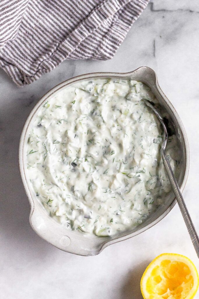 Bowl of homemade tzatziki with a spoon in it. Next to it is a kitchen towel and half a juiced lemon.