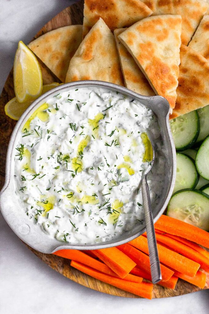 Large bowl of tzatziki sauce with a spoon coming out of it and topped with fresh dill and olive oil. Around it is pita bread, cucumber slices, sliced carrots, and lemon wedges.