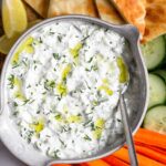 Tzatziki sauce garnished with dill and olive in a large bowl with a spoon in it. It is surrounded by sliced pita bread, sliced cucumber, carrot sticks, and lemon wedges.
