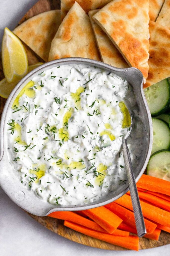 Tzatziki sauce garnished with dill and olive in a large bowl with a spoon in it. It is surrounded by sliced pita bread, sliced cucumber, carrot sticks, and lemon wedges.