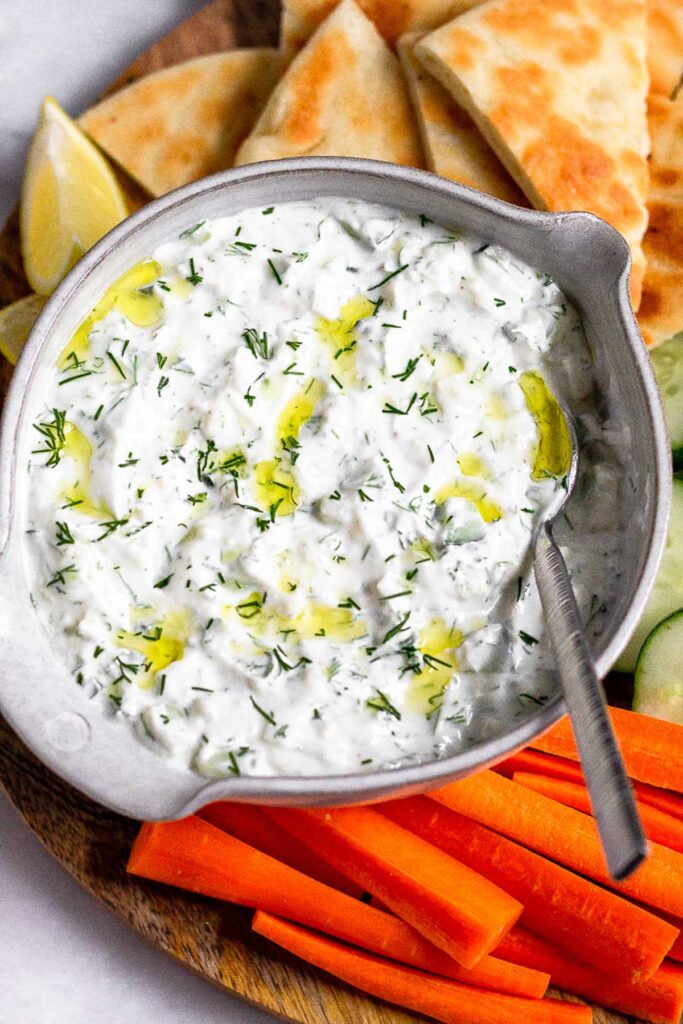 Close up of a bowl of tzatziki sauce garnished with dill and olive oil with a spoon in it. Around it is chopped carrots, cucumbers, and pita bread for dipping.