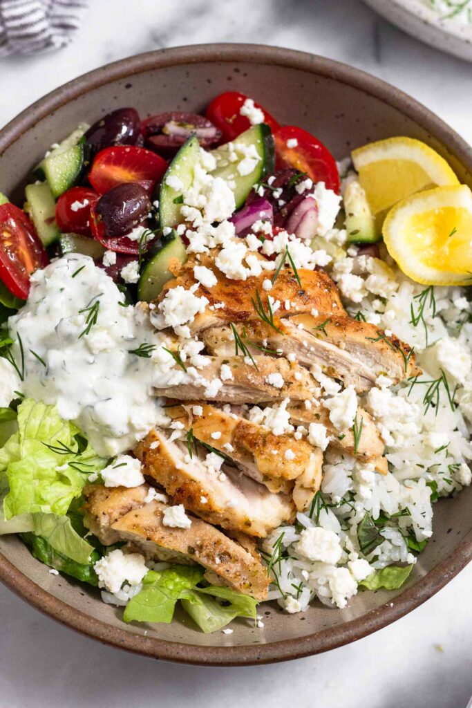 Bowl filled with chopped lettuce, tomatoes, cucumber, and olives, white rice, sliced chicken thighs, tzatziki, two lemon wedges and topped with feta. It is garnished fresh dill.