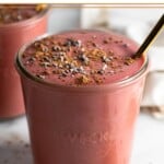 Beet smoothie in a jar topped with cacao nibs and a straw in it. In front of it is a teaspoon filled with cacao powder. Behind it is another jar filled with smoothie.