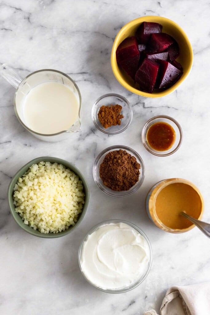 White marble counter with a bowl of diced beets, a bowl of cinnamon, a bowl of cacao powder, a bowl of honey, a bowl of cashew butter, a bowl of greek yogurt, a bowl of frozen cauliflower rice, and a jar of milk.