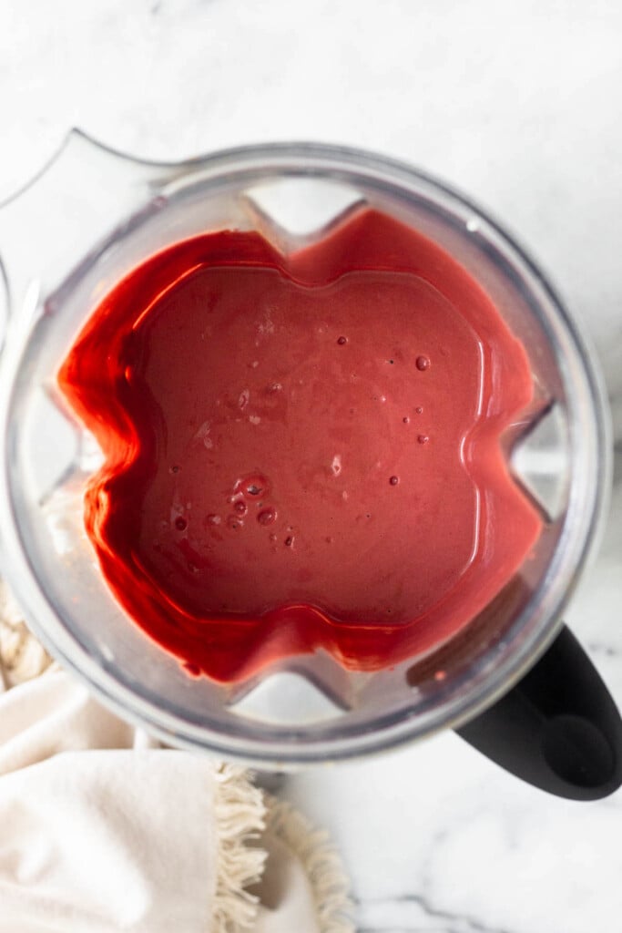 Reddish pink smoothie in a blender with a cream colored towel next to it.