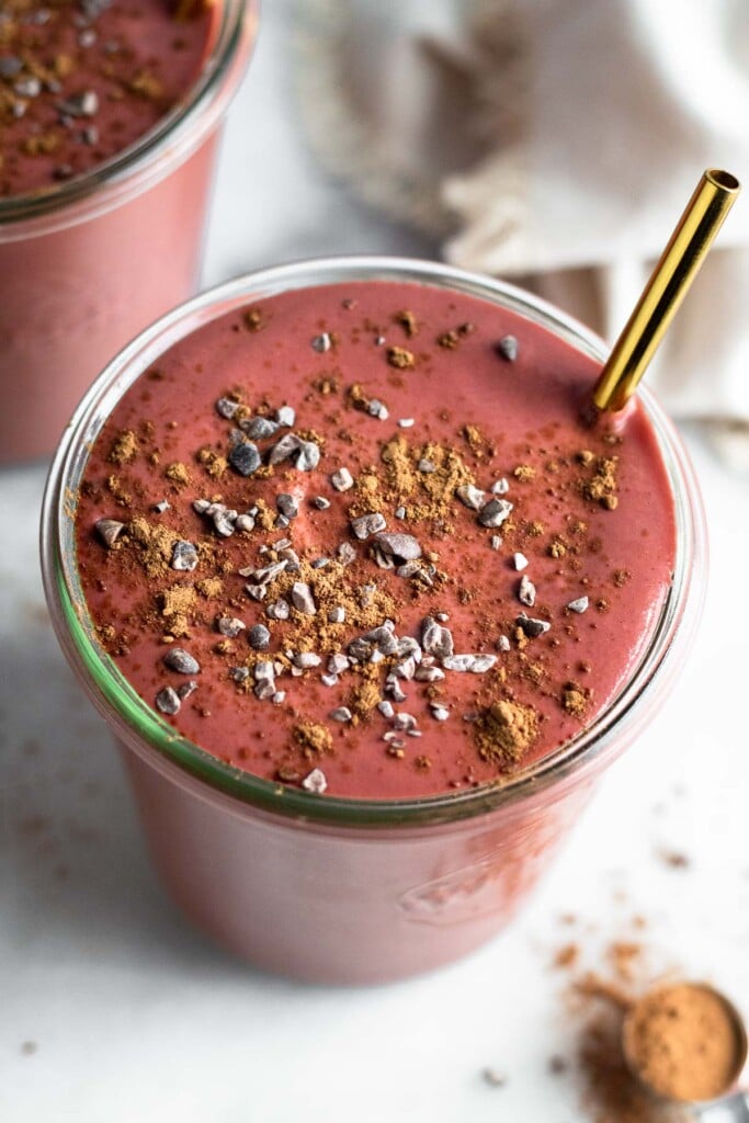 Overhead shot of a beet smoothie topped with cacao nibs and cacao powder. A gold straw is also in it. Next to is another smoothie and a teaspoon filled with cacao powder.