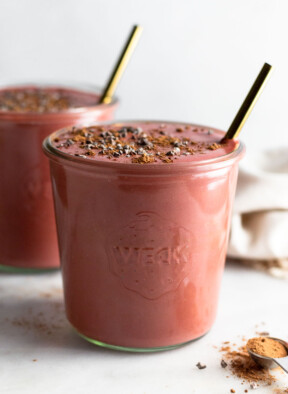 Beet smoothie in a jar topped with cacao nibs and a straw in it. In front of it is a teaspoon filled with cacao powder. Behind it is another jar filled with smoothie.