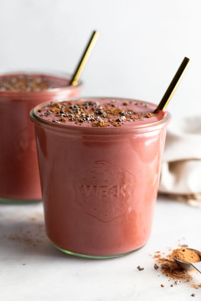 Beet smoothie in a jar topped with cacao nibs and a straw in it. In front of it is a teaspoon filled with cacao powder. Behind it is another jar filled with smoothie.