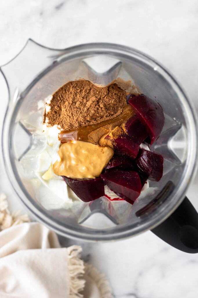 Blender filled with milk, yogurt, cauliflower rice, beets, cashew butter, cacao powder, chocolate protein powder, and cinnamon.