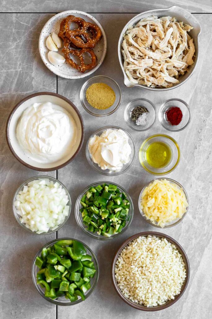 Grey marble counter top with a bowl of shredded chicken, three small bowls of spices, a bowl of olive oil, a bowl of shredded cheese, a bowl of cauliflower rice, a bowl of diced bell pepper, a bowl of diced jalapeños, a bowl of diced onions, a bowl of cream cheese, a bowl of greek yogurt, and a plate with some pretzels and garlic cloves on it.