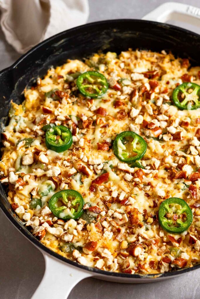 Jalapeno chicken casserole topped with melted cheese, crushed pretzels, and a sliced jalapeños in a large cast iron skillet.