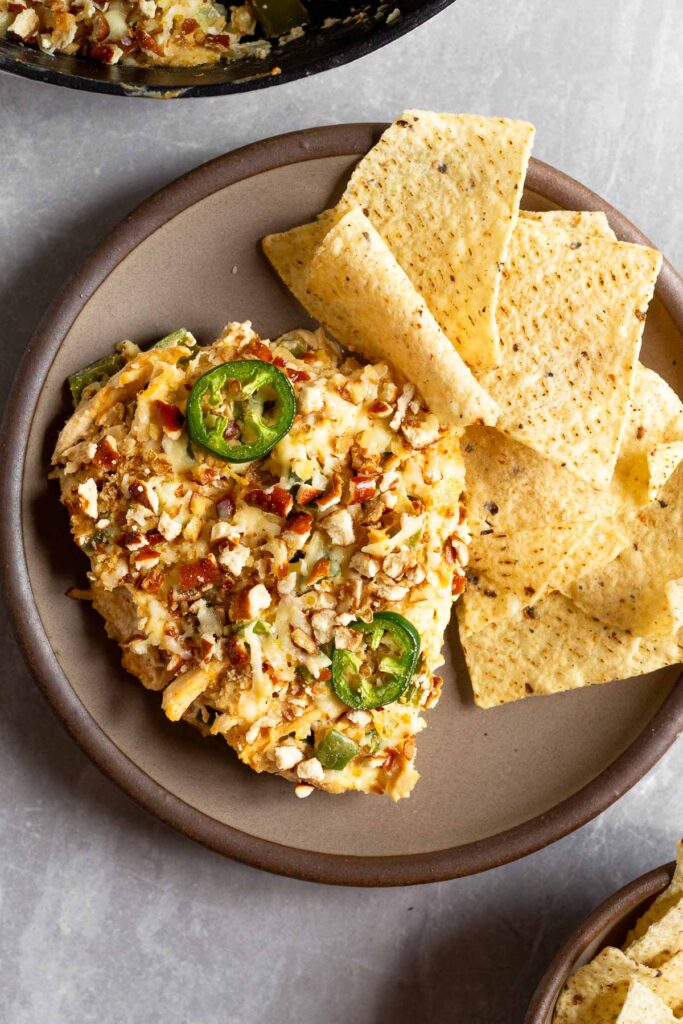 A plate with a piece of jalapeño chicken casserole and a handful of chips on it. Off to the side is a dish with more casserole in it and bowl of chips.