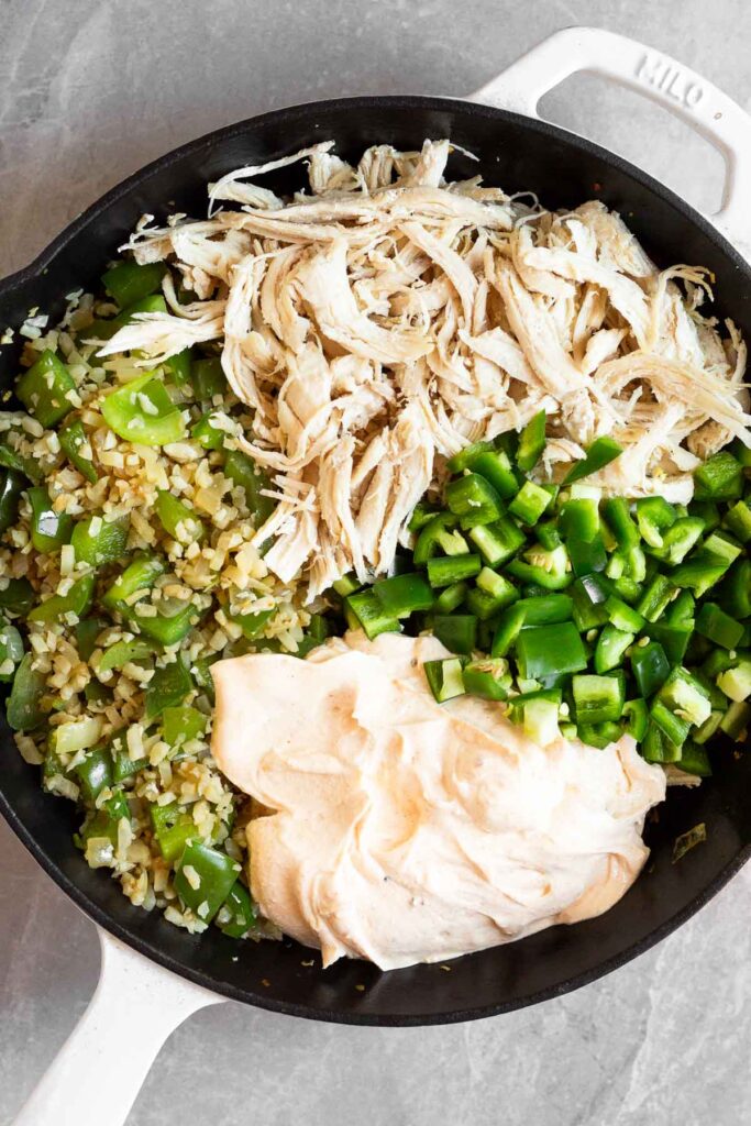 Large cast iron skillet filled with a pile of sautéed veggies, a pile of shredded chicken, a pile of diced jalapeños, and a pile of a cream cheese yogurt mixture.
