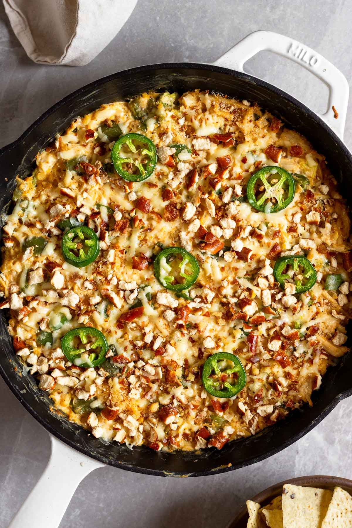 Cast iron skillet filled with jalapeño popper chicken casserole topped with melted cheese, crumbled pretzels, and sliced jalapeños. Next to it is a kitchen towel and bowl of chips.