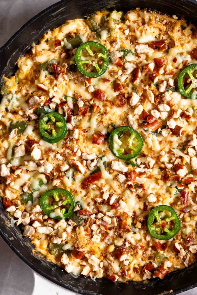 Close up of chicken jalapeño popper casserole topped with melted cheese, crushed pretzels, and sliced jalapeños in a large cast iron skillet.