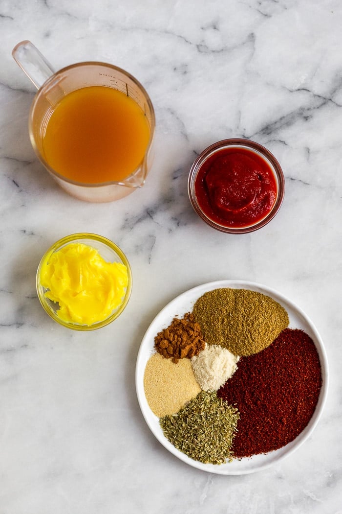 A white counter filled with a plate of spices, a bowl of ghee, bowl of tomato pasta, and a cup filled with broth.