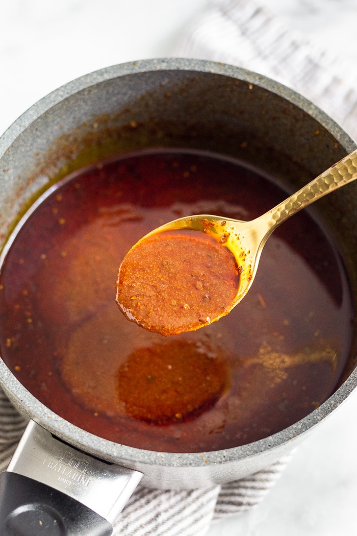Pot filled with homemade enchilada sauce with a spoonful hovering of it. The focus is on the spoon.